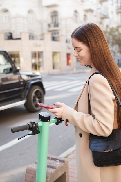 Photo verticale d'une femme élégante et joyeuse uaing application mobile pour louer un scooter électrique dans la ville