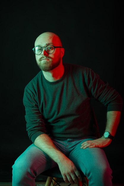 Photo verticale d'un homme chauve barbu assis sur une chaise sur fond noir Studio tourné avec lumière rouge et verte