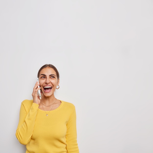 Photo verticale d'une jeune femme joyeuse et heureuse a une conversation amusante via un smartphone qui rigole discute positivement de quelque chose de drôle porte un pull jaune isolé sur fond blanc espace de copie vierge