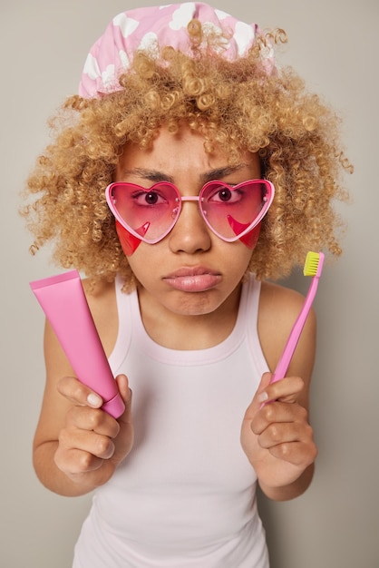 La photo verticale d'une jeune femme mécontente regarde avec colère les lèvres des sacs à main applique des patchs d'hydrogel sous les yeux tient le dentifrice et la brosse à dents prend soin des dents et du visage isolés sur fond gris