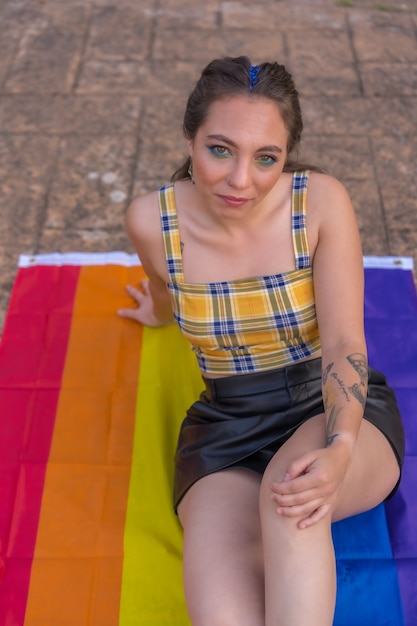 Une photo verticale d'une jeune femme souriante de race blanche assise sur un drapeau de fierté LGBT à l'extérieur