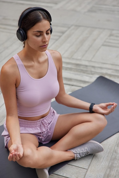 Photo verticale d'une jeune femme sportive paisible assise dans une pose de lotus médite sur un tapis de fitness respire profondément garde les yeux fermés écoute de la musique relaxante via des écouteurs essaie de trouver l'harmonie Reste calme