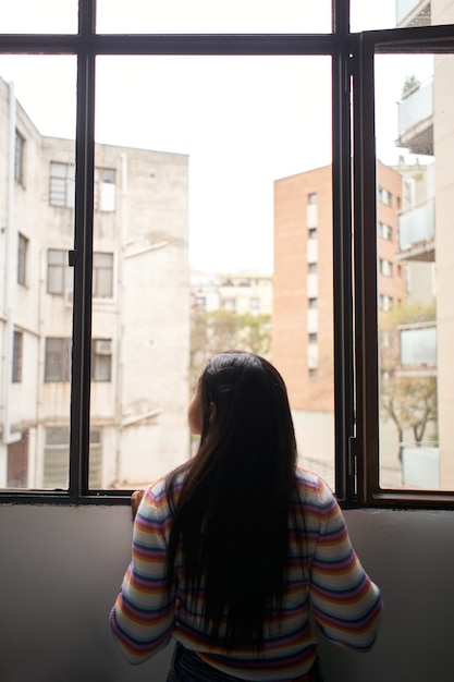 photo verticale. Vue arrière fille africaine se penchant par la fenêtre, regardant à l'extérieur où il y a de grands bâtiments.