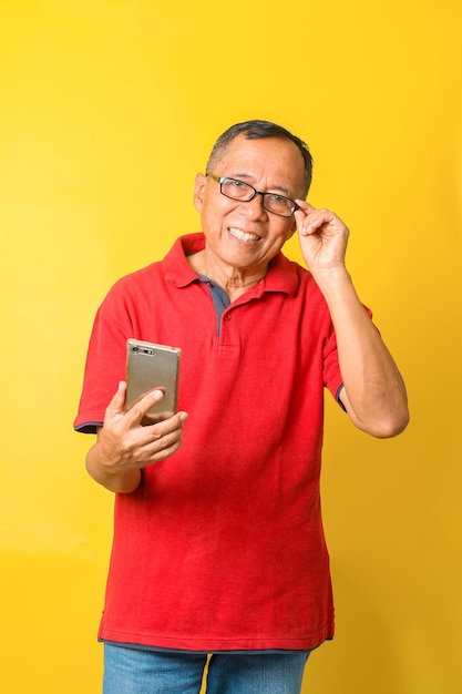 Photo d'un vieil homme asiatique souriant et tenant un smartphone et des lunettes portant un t-shirt rouge isolé o