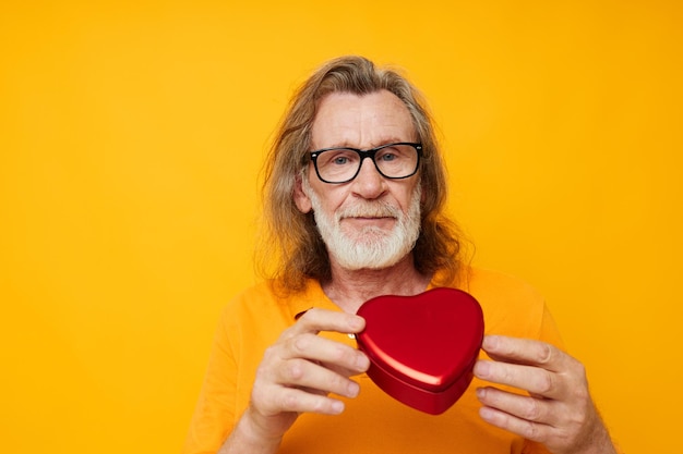 Photo d'un vieil homme à la retraite t-shirt jaune et lunettes posant une boîte en forme de coeur sur fond jaune