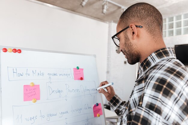 Photo vue arrière d'un jeune homme d'affaires africain portant des lunettes au bureau travaillant avec un tableau à feuilles mobiles.