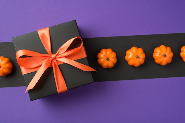 Photo vue de dessus de la boîte-cadeau noire de composition d'halloween avec noeud de ruban orange et petites citrouilles d'affilée sur fond isolé et violet avec fond