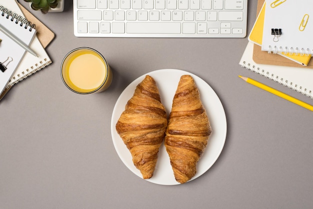 Photo vue de dessus des cahiers de clavier en milieu de travail pinces à reliure stylo crayon jaune pot de fleurs verre de jus et assiette avec deux croissants sur fond gris isolé