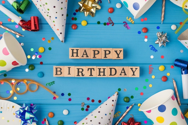 Photo vue de dessus de la composition d'anniversaire cubes en bois étiquetés joyeux anniversaire au milieu des gobelets en papier à pois et des assiettes blanches accessoires de fête sur fond de table en bois bleu isolé