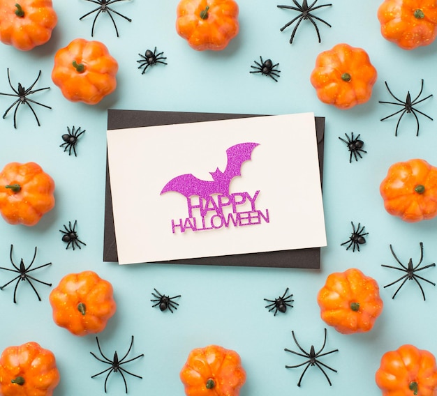 Photo vue de dessus des décorations d'halloween enveloppe noire chauve-souris scintillante et inscription joyeux halloween sur carte blanche citrouilles et araignées sur fond bleu pastel isolé