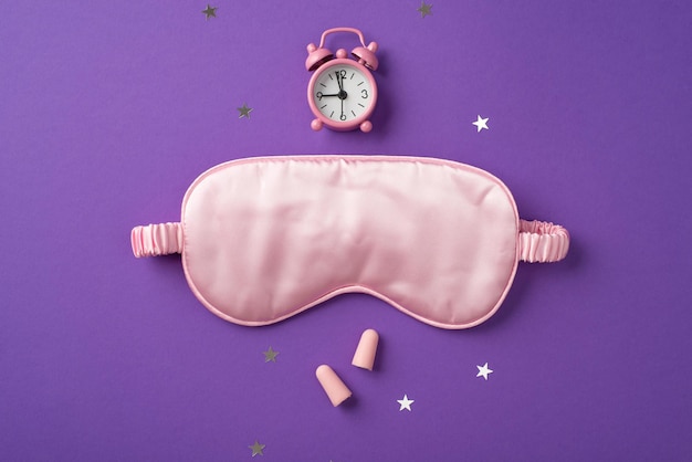 Photo vue de dessus du grand masque de sommeil en soie et de la petite horloge rose au-dessus avec des confettis dispersés en forme d'étoiles et deux bouchons d'oreilles roses sur fond violet