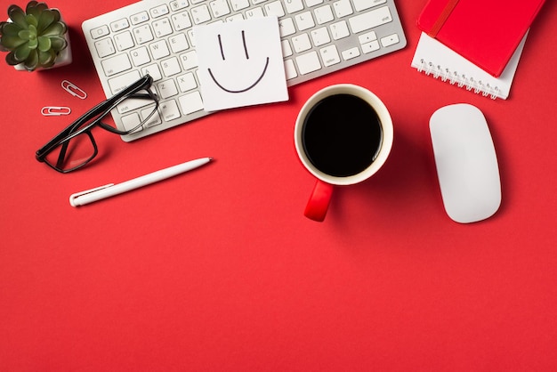 Photo vue de dessus du papier à lettres autocollant avec visage souriant dessiné sur clavier blanc souris plante verres clips stylo tasse de café rouge et deux cahiers sur fond rouge vif isolé avec espace de copie