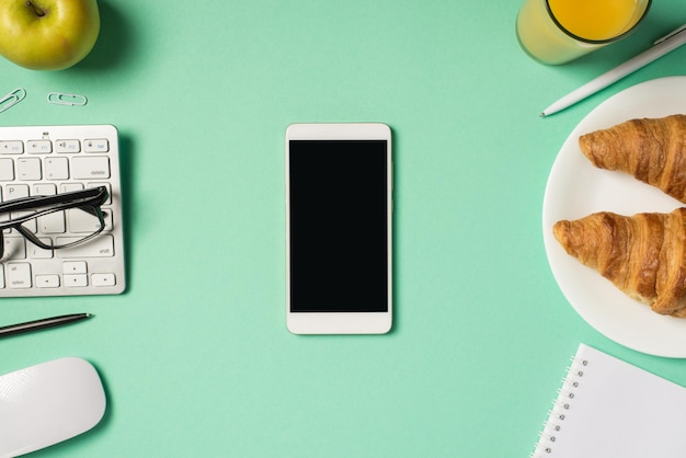 Photo vue de dessus du smartphone sur le lieu de travail au milieu clavier lunettes souris stylos clips cahier pomme verre de boisson et assiette avec deux croissants sur fond turquoise isolé avec fond