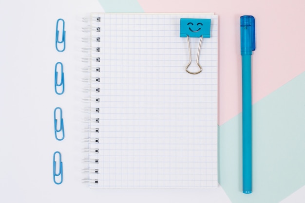 Photo vue de dessus des fournitures scolaires. Ensemble de cahier à spirale ouvert, stylo, trombones sur fond rose et bleu