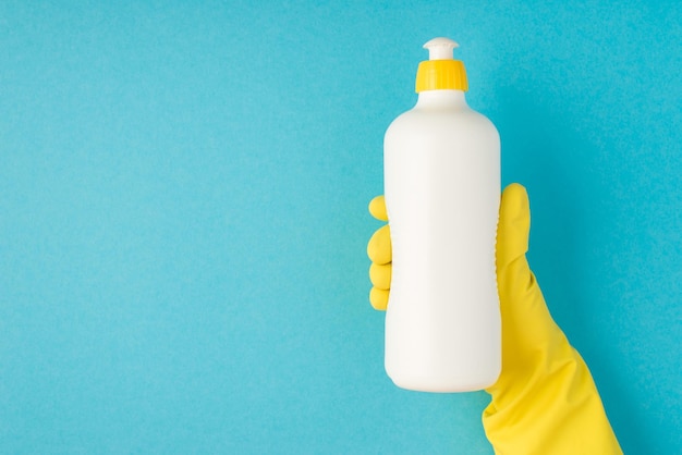 Photo vue de dessus de la main dans un gant en caoutchouc jaune tenant une bouteille de mousse détergente blanche sans étiquette sur fond bleu pastel isolé avec espace vide