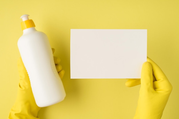 Photo photo vue de dessus des mains dans des gants en caoutchouc jaune tenant une bouteille de détergent blanc sans étiquette et une carte en papier blanc sur fond jaune isolé avec espace vide