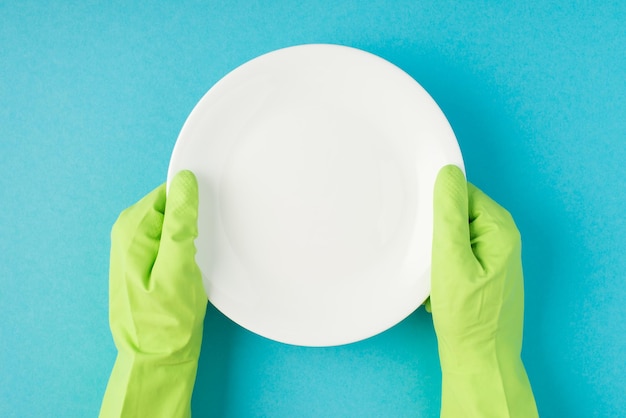 Photo vue de dessus des mains dans des gants en caoutchouc vert tenant une assiette propre blanche sur fond bleu pastel isolé avec espace pour copie