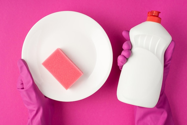 Photo vue de dessus des mains dans des gants roses tenant un tampon à récurer rose sur un plat blanc propre et une bouteille de gel détergent blanc sur fond rose isolé avec fond