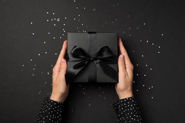 Photo vue de dessus à la première personne des mains en chemise à pois tenant une boîte-cadeau noire avec un nœud en ruban de satin noir sur des paillettes brillantes sur fond noir isolé