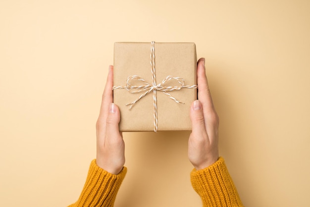 Photo photo vue de dessus à la première personne des mains dans un chandail orange tenant une boîte-cadeau en papier kraft avec un arc de ficelle sur fond beige isolé