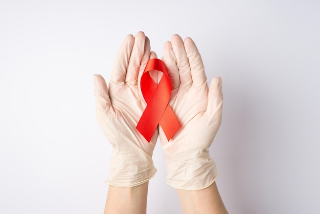 Photo vue de dessus à la première personne des mains dans des gants de protection médicale tenant un ruban rouge dans les paumes symbole de la sensibilisation au sida sur fond blanc isolé