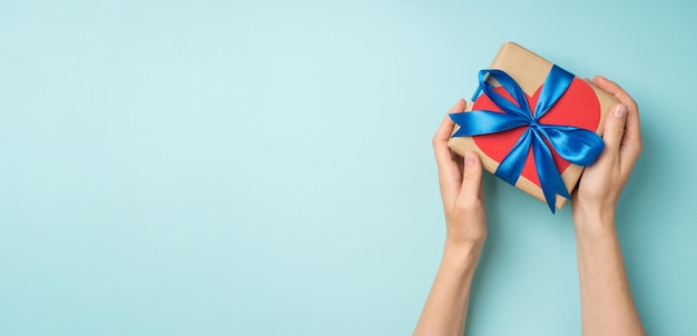 Photo vue de dessus à la première personne des mains donnant une boîte-cadeau en papier kraft élégant avec un arc en ruban de satin bleu et un coeur en papier rouge sur fond bleu pastel isolé avec un espace vide