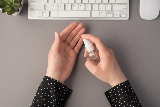 Photo vue de dessus à la première personne de mains féminines à l'aide d'un flacon pulvérisateur de désinfectant clavier blanc souris et plante sur fond gris isolé