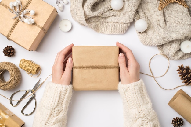 Photo vue de dessus à la première personne de mains féminines dans un chandail enveloppant une boîte-cadeau en papier kraft avec une écharpe tricotée en ficelle des boules de sapin de Noël et des outils pour l'artisanat sur fond blanc isolé