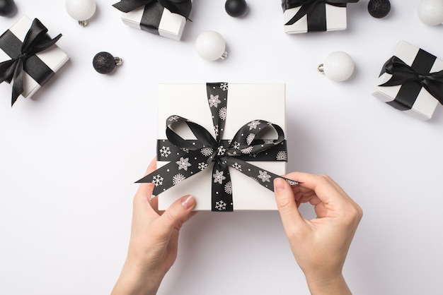 Photo vue de dessus à la première personne de mains féminines déballant une boîte-cadeau blanche avec un arc de ruban noir présente des boules de sapin de Noël blanches et noires sur fond blanc isolé