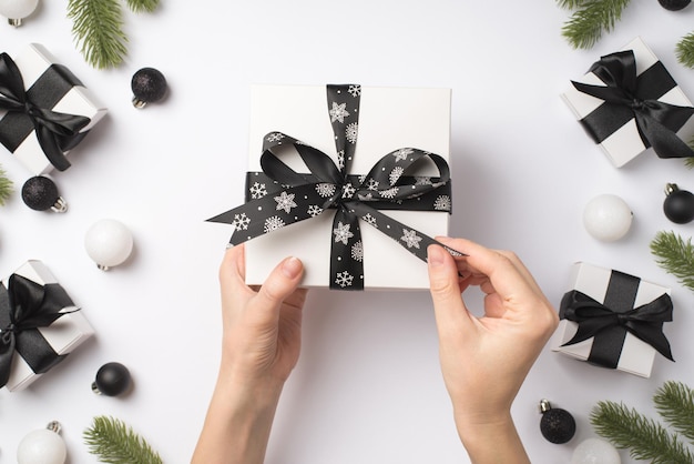 Photo vue de dessus à la première personne de mains féminines déballant une boîte-cadeau blanche avec un noeud de ruban noir sur des décorations de Noël blanches et noires, des branches de pin, des cadeaux et des boules sur fond blanc isolé