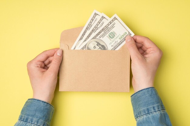 Photo vue de dessus à la première personne de mains féminines tenant une enveloppe en papier kraft ouverte avec des billets de cent dollars sur fond jaune isolé avec espace pour copie