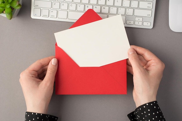 Photo vue de dessus à la première personne de mains féminines tenant une enveloppe rouge ouverte avec une carte blanche clavier blanc souris et plante sur fond gris isolé avec espace de copie