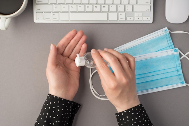 Photo vue de dessus à la première personne des mains de la femme à l'aide d'une bouteille transparente de désinfectant deux masques médicaux clavier blanc souris et tasse de café sur fond gris isolé