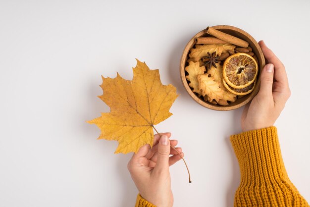 Photo vue de dessus à la première personne de mains de femme en pull jaune tenant une feuille d'érable d'automne jaune et un bol en bois avec des tranches de citron séchées anis et des bâtons de cannelle sur fond blanc isolé