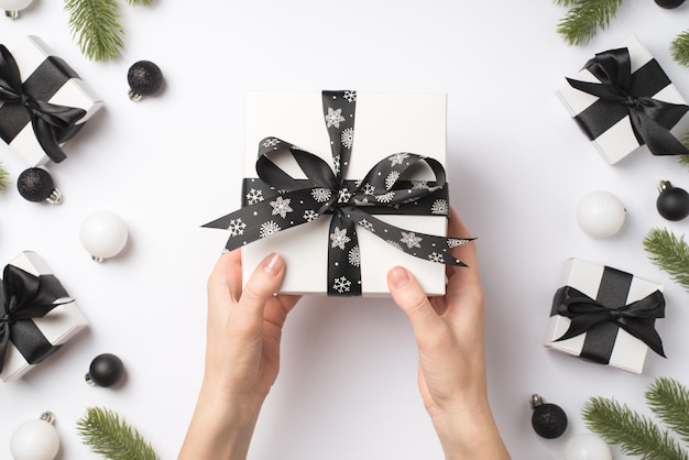 Photo vue de dessus à la première personne des mains d'une jeune femme tenant une boîte-cadeau blanche avec un noeud de ruban noir sur des décorations de Noël blanches et noires, des brindilles de pin, des cadeaux et des boules sur fond blanc isolé