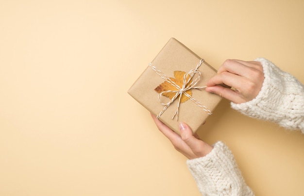 Photo vue de dessus à la première personne des mains d'une jeune fille en pull blanc déballant une boîte-cadeau en papier kraft avec un arc en ficelle et une feuille d'automne jaune sur fond beige isolé avec un espace vide