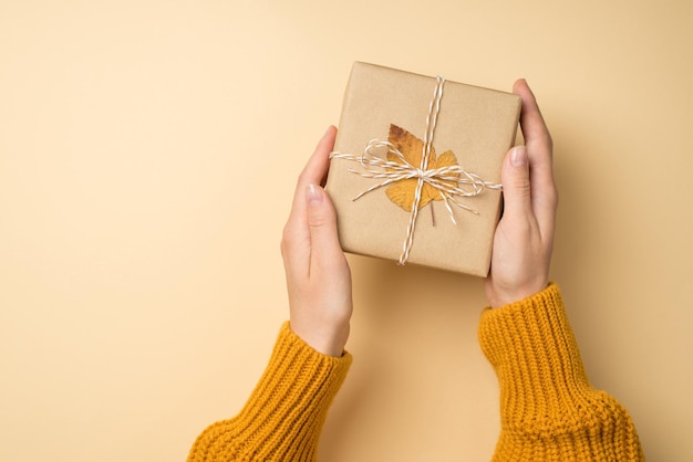 Photo vue de dessus à la première personne des mains d'une jeune fille en pull tricoté jaune tenant une boîte-cadeau en papier kraft avec un arc en ficelle et une feuille d'automne jaune sur fond beige isolé avec fond