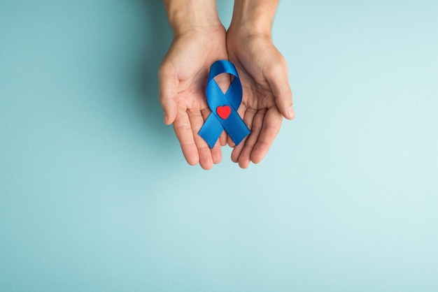Photo vue de dessus à la première personne des mains masculines démontrant un ruban bleu avec un petit coeur rouge dans les paumes symbole de la sensibilisation au cancer de la prostate sur fond bleu pastel isolé avec espace vide