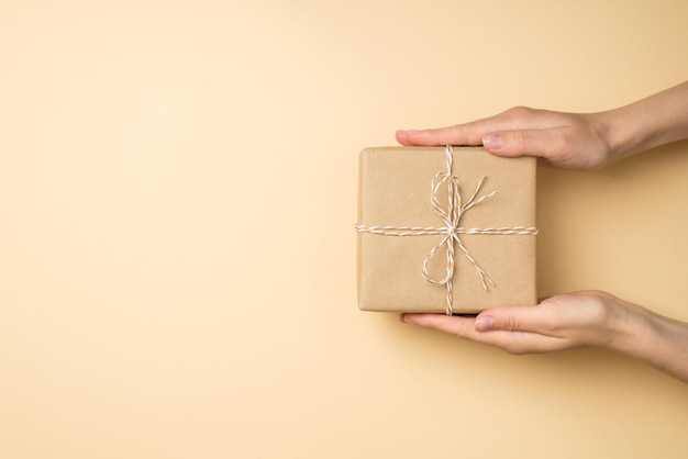 Photo photo vue de dessus à la première personne des mains tenant une boîte-cadeau en papier kraft avec noeud de ficelle sur fond beige isolé avec espace pour copie