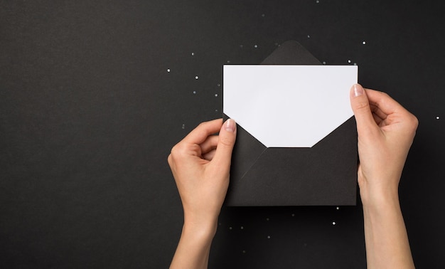 Photo vue de dessus à la première personne des mains tenant une enveloppe ouverte noire avec une carte blanche sur des paillettes sur fond noir isolé avec un espace vide