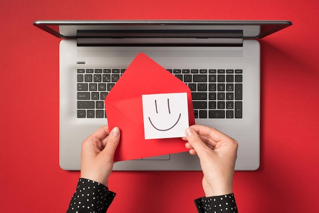 Photo vue de dessus à la première personne des mains tenant une enveloppe rouge vif et un papier autocollant blanc avec un visage souriant dessiné sur un ordinateur portable gris ouvert sur fond rouge isolé