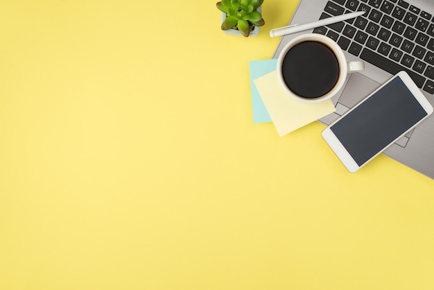 Photo vue de dessus d'une tasse de boisson autocollant jaune et bleu note papier stylo smartphone sur ordinateur portable et pot de fleurs sur fond jaune pastel isolé avec espace vide