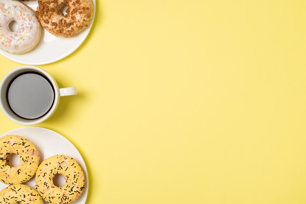 Photo vue de dessus d'une tasse de café et de deux assiettes avec des beignets glacés de couleur différente sur fond jaune pastel isolé avec espace pour copie