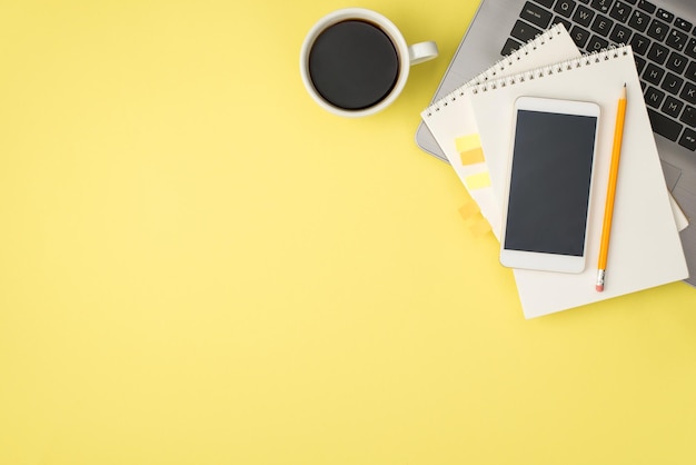 Photo vue de dessus d'une tasse de crayon à café pour ordinateur portable et d'un smartphone sur deux cahiers en spirale avec des signets adhésifs en plastique jaune sur fond jaune pastel isolé avec espace vide