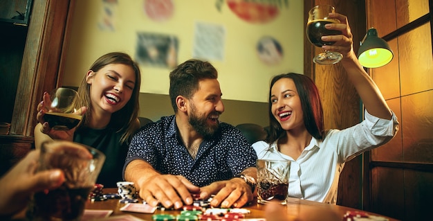 Photo vue latérale d'amis masculins et féminins assis à une table en bois. Hommes et femmes jouant au jeu de cartes. Mains avec gros plan d'alcool. Concept de poker, de divertissement en soirée et d'excitation