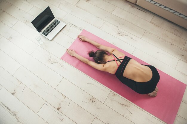 Photo vue supérieure d'une femme de race blanche pratiquant le yoga à la maison portant des vêtements de sport et à l'aide d'un ordinateur portable sur le sol