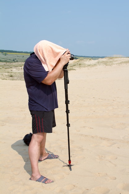 Un photographe adulte avec une serviette sur la tête provenant de la chaleur prend des photos avec une caméra sur un support dans le désert