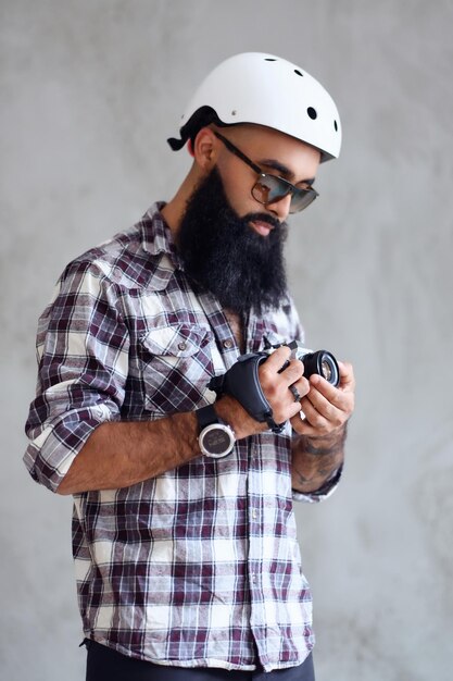Un photographe amateur hipster barbu avec des tatouages sur les bras, vêtu d'une chemise polaire tient un appareil photo reflex numérique compact sur fond gris.