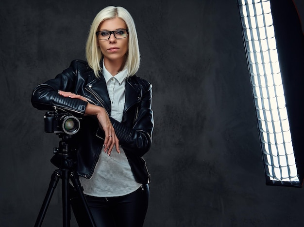 Une photographe blonde vêtue d'une veste en cuir noir tient un appareil photo numérique professionnel et un trépied.