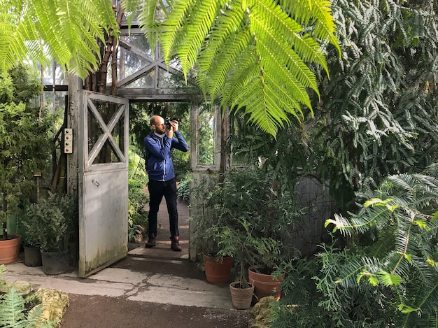 Photographe homme barbu prenant une photo à la caméra dans une serre, debout avec une vieille porte en acier ouverte sur une journée ensoleillée.